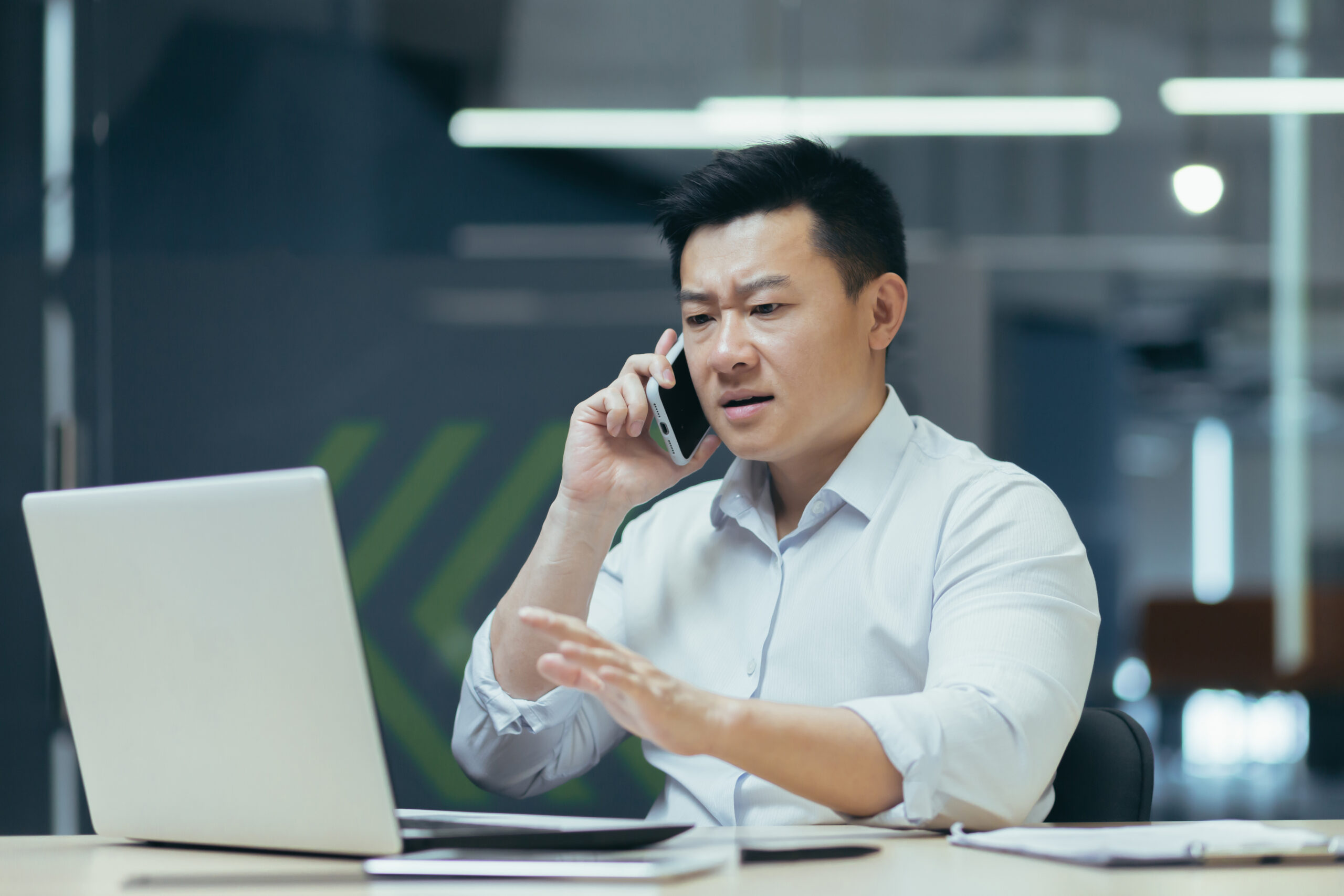 Worried and nervous young Asian man talking on the phone in the office, looking at the laptop, solving problems, questions